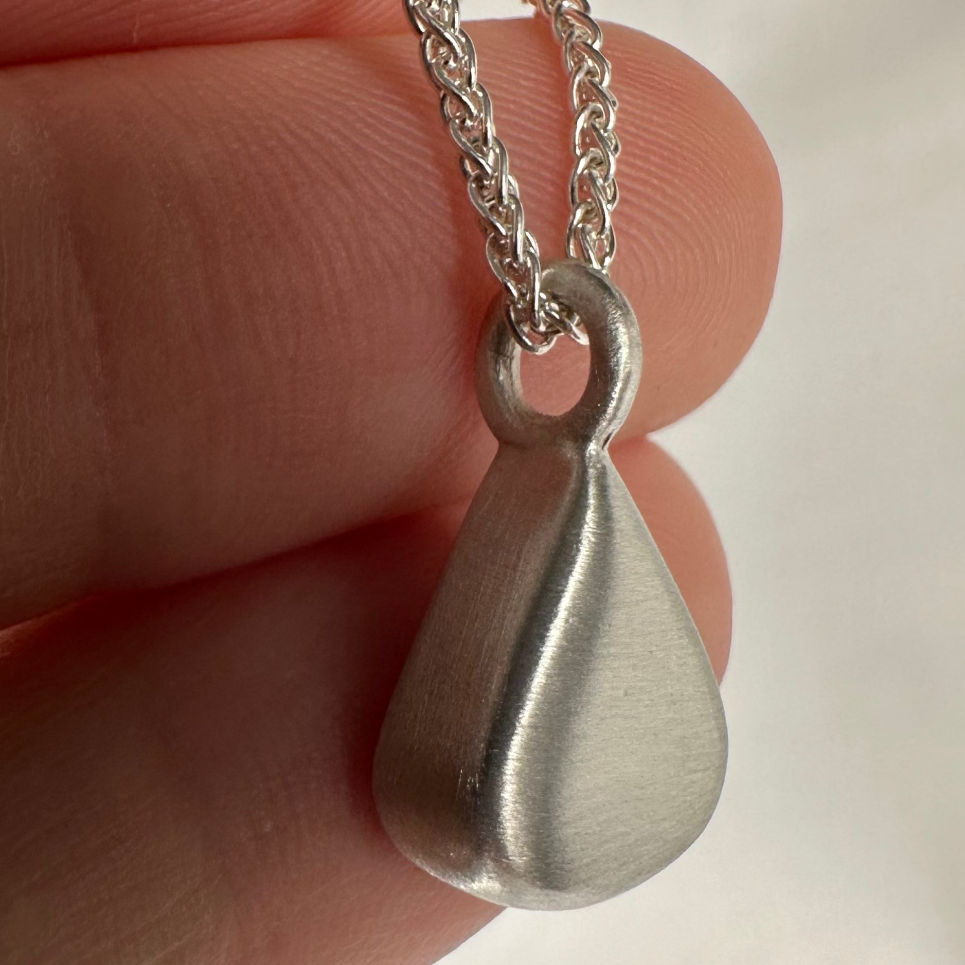 Silver teardrop pendant on a chain held between fingers against a neutral background