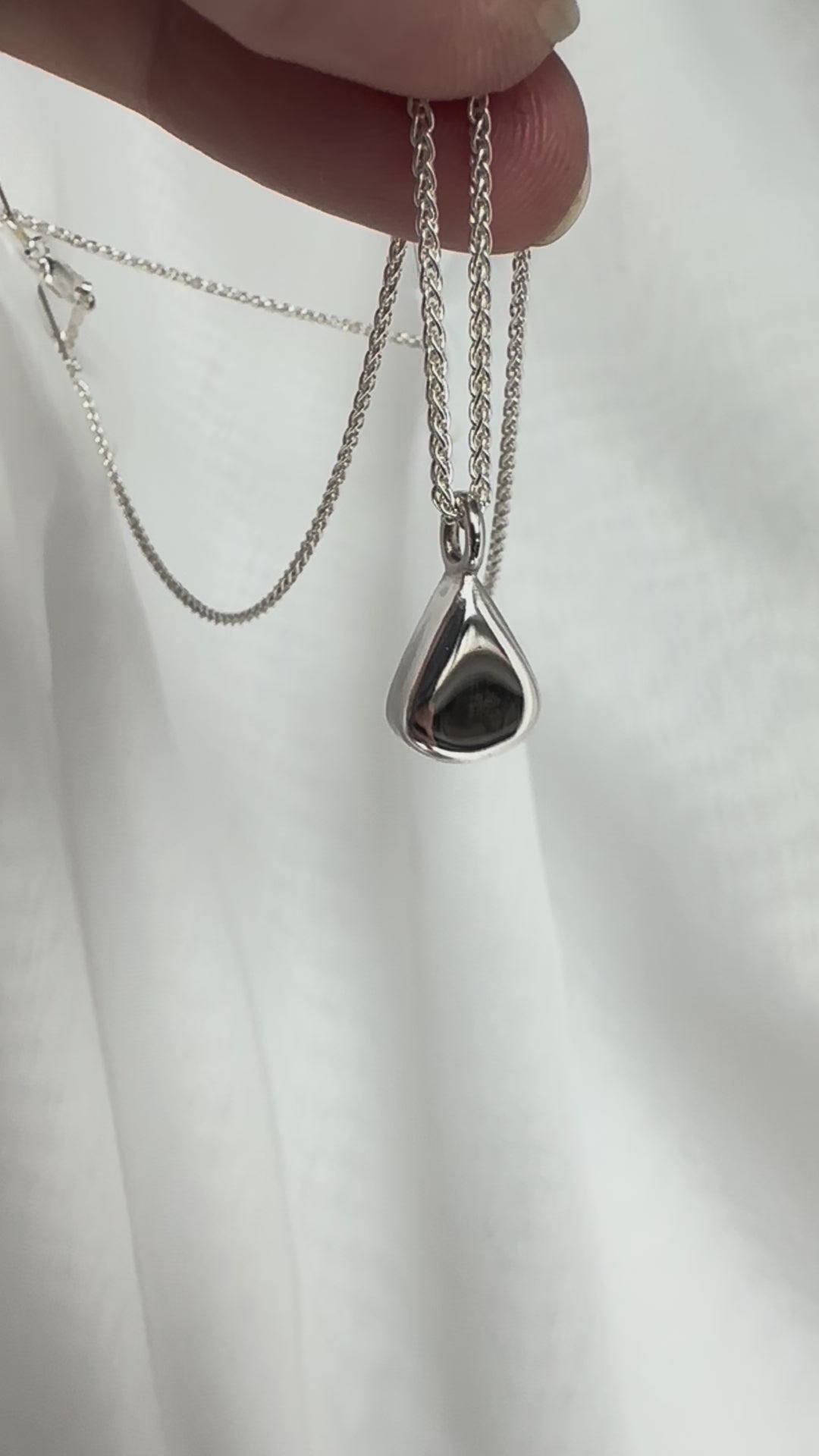 Sterling Silver Pear Pendant on a spiga chain being held between fingers in front of a white fabric backdrop, being waved gently to catch the light on its mirror shine.