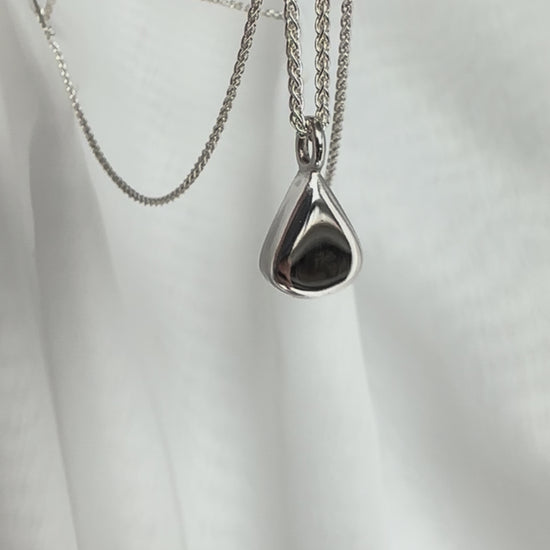 Sterling Silver Pear Pendant on a spiga chain being held between fingers in front of a white fabric backdrop, being waved gently to catch the light on its mirror shine.
